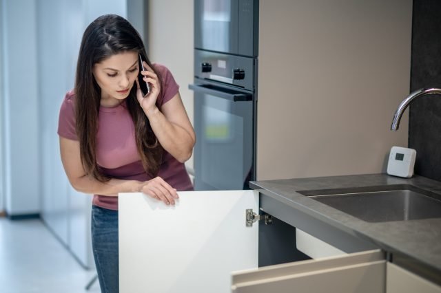 Problem. Young woman with long dark hair looking unpleasantly surprised looking under kitchen sink talking on smartphone at home